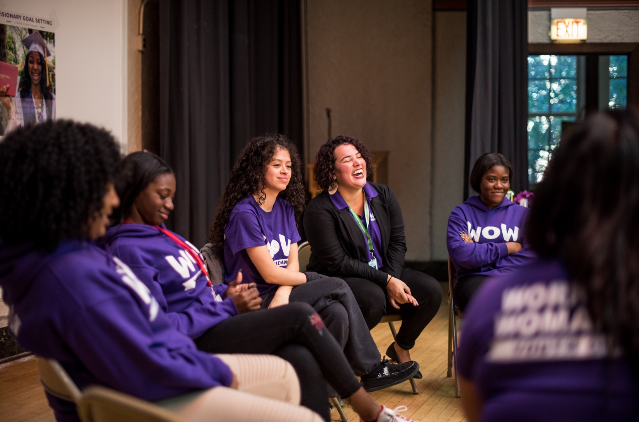 An image of WOW women sitting in a circle laughing and wearing matching purple t-shirts inside a room.