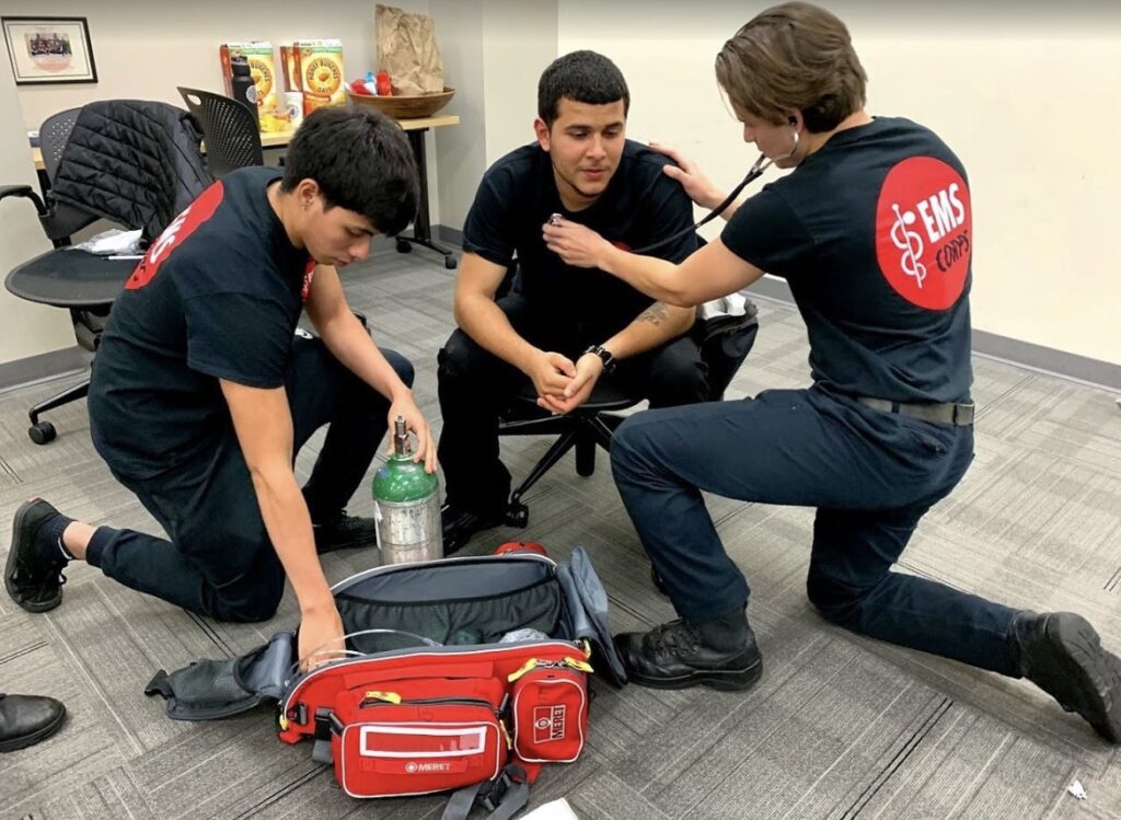 EMS Corps participants in Newark sitting on the ground practicing their skills with an oxygen tank and stethoscope.