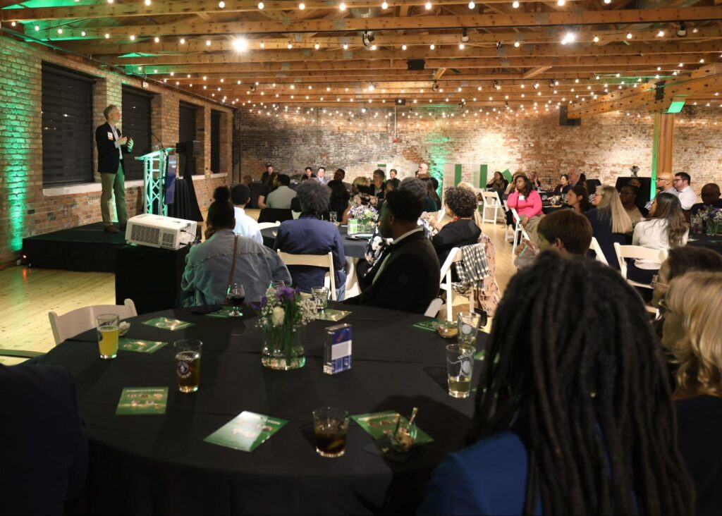 HomeStart’s President, Matthew Pritchard stands on stage speaking to a room illuminated with string lights and filled with individuals at black circular tables attending the Cinnovation event.