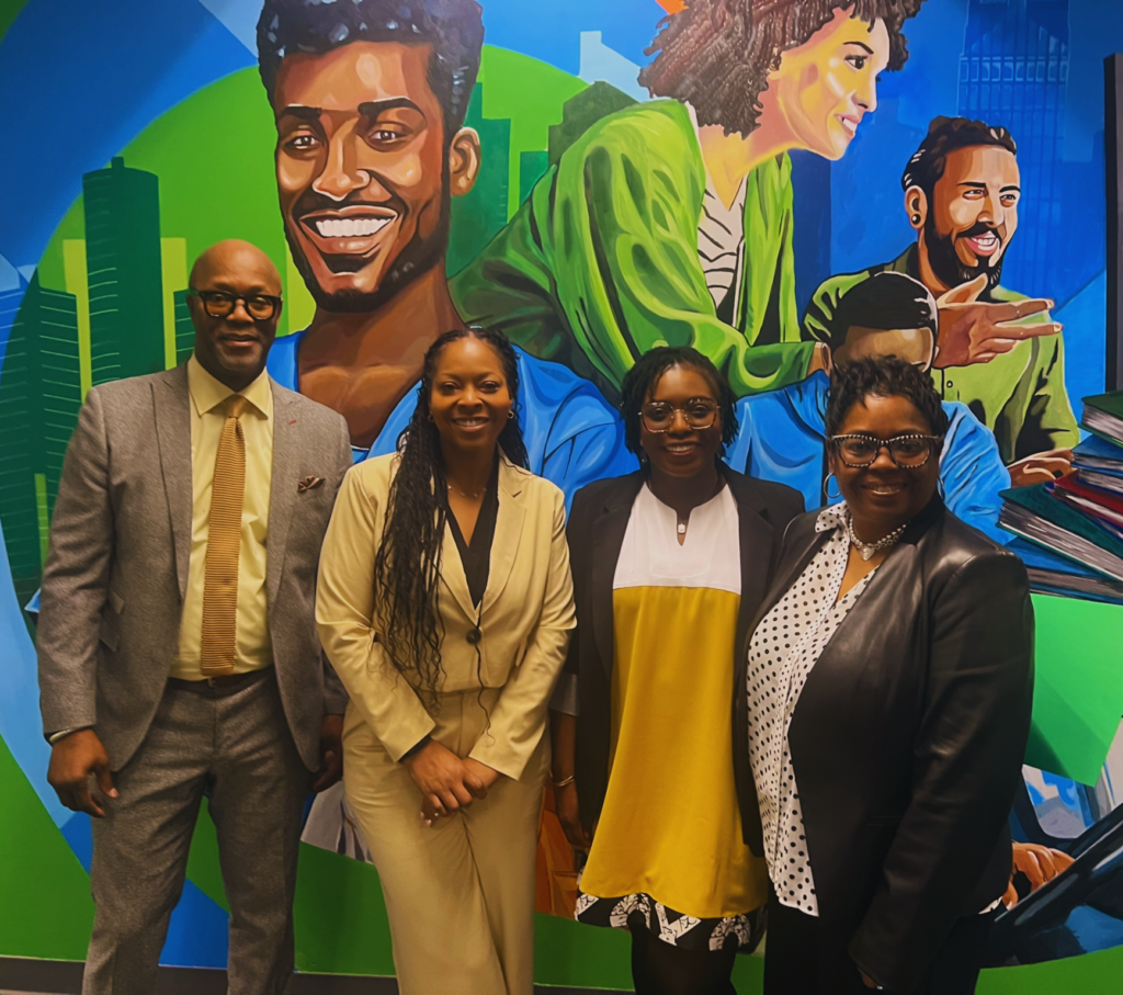A group of four people stand in front of a mural portraying several career paths offered by Center for Employment Opportunities in the city of Detroit. From left to right, the image includes Terrell Topps, Margrit Allen, Olu Martins, and Nina Hicks