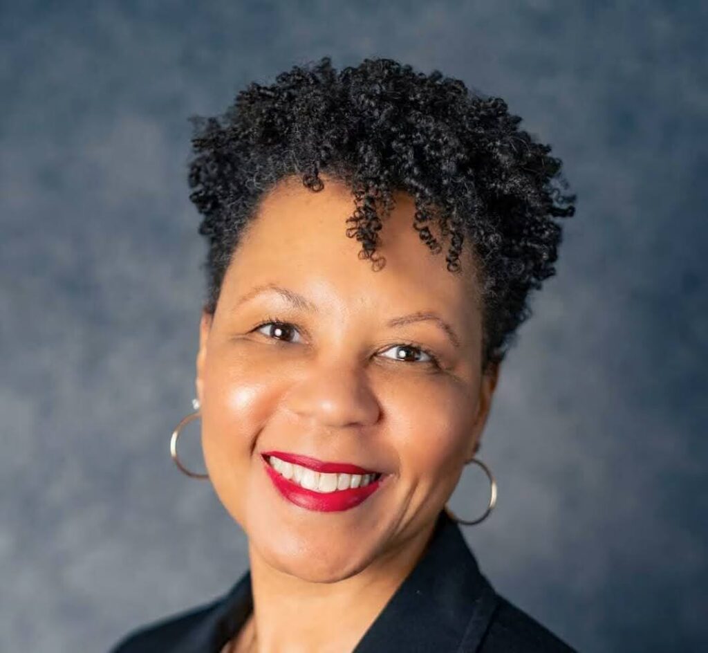 Headshot image of Dr. Kelly Brittain, a Black woman with short hair wearing a black blazer over a white shirt with gold earrings and red lipstick